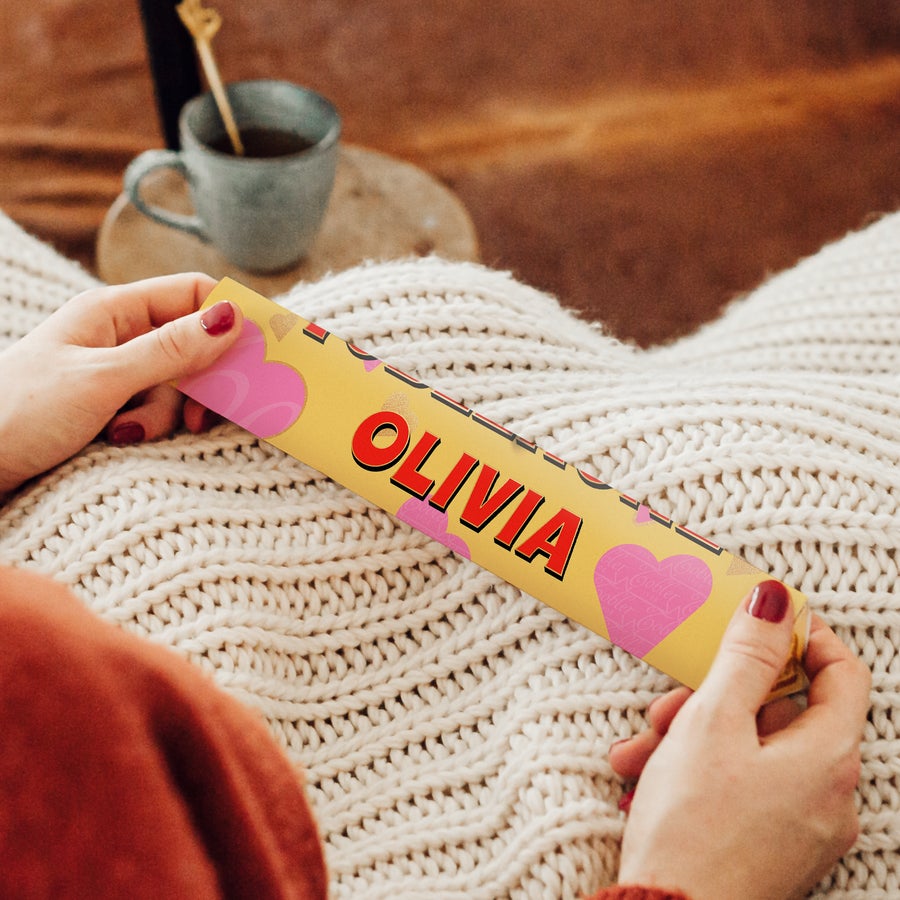 Personalised Toblerone Chocolate Bar - Love Hands holding a personalized Toblerone bar with the name Olivia printed on a yellow wrapper, featuring pink heart designs